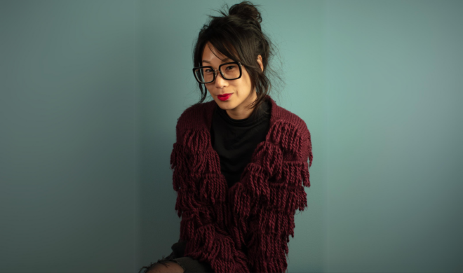 A photograph portrait of artist Annie Wong. She is an Asian woman with fair skin, dark hair tied into a bun, and dark rimmed glasses. She is wearing a black top and red fringed cardigan. She is looking into the camera. She is wearing red lipstick.