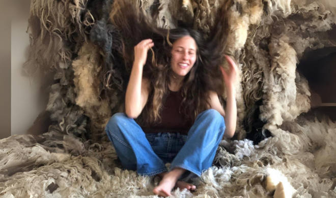 A photograph of dancer Angela Vitovec. She is white, with long brown hair. She is sitting inside a furry cave. She is wearing a brown top and blue jeans, and is barefoot. There is a smile on her face.