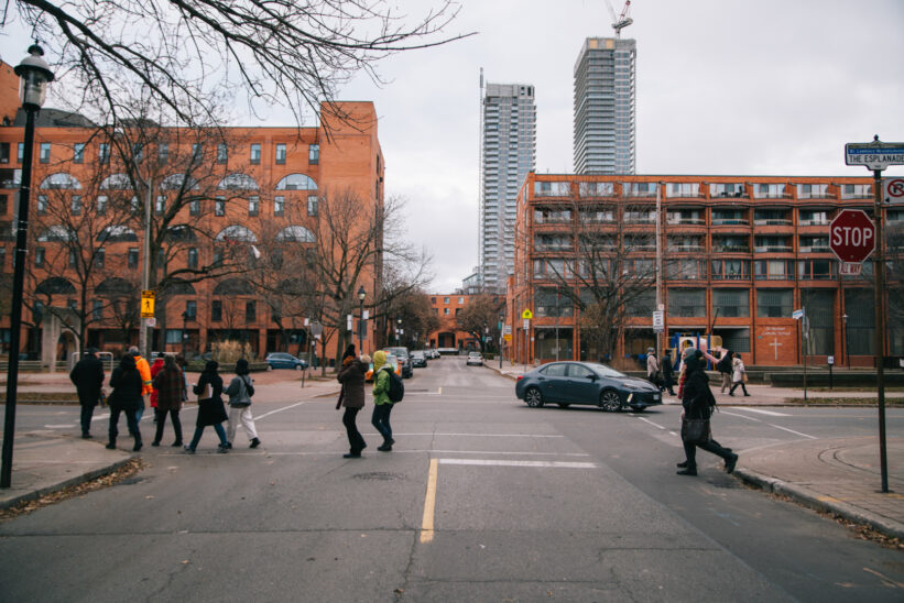 A photograph of a group of people walking across the street together on The Esplanade in Toronto. Buildings and condos can be seen in the background.
