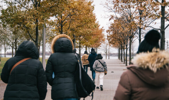 A photograph of a group of people walking together down a tree-lined dock. They are wearing winter jackets.