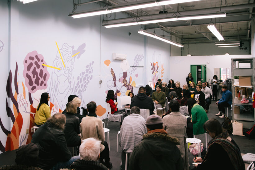 A photo of a group of at least 40 people in a room, sitting in a large group and circle, listening to a smaller group of 6 people speaking. The room has a large mural on one wall.