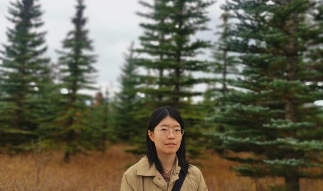 A photo portrait of Jingshu Yao. She is a Chinese woman with mid-length black hair, brown eyes and square-shaped metal wire glasses. She is wearing a beige trench coat in front of a roll of trees.
