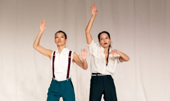 Justine A. Chambers, a mixed-race Black woman, and Laurie Young, an Asian woman, stand against a white curtain backdrop. Justine is wearing a white dress shirt and dark slacks. Laurie is wearing a sleeveless white dress shirt, suspenders, and blue slacks. Both of them have their arms raised, one arm reaching up over the other. Their gazes are wayward.