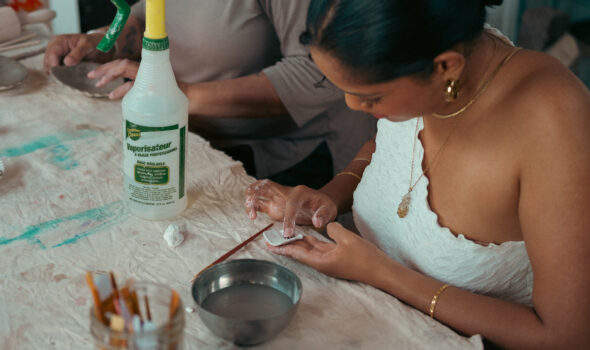 A photo of a person with brown skin, dark hair pushed back, a white top, and gold jewellery working with a small piece of soft clay in their hands. The table they are working at has a tattered cloth, spray bottle, bowl of water, and paint brushes.
