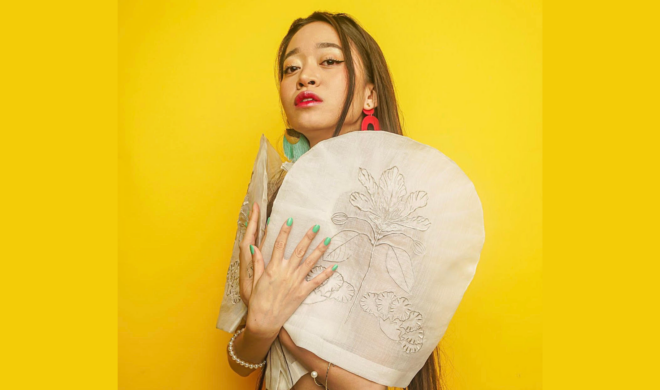 A photo portrait of Shadel Chavez. She is Filioina-Canadian. She is against a bright yellow backdrop. She is wearing a beige tereno-styled top with puffed shoulders with embroidered designs, with her hands crossed over one another to hold the sleeves. Her nail polish is teal. She is wearing pink and teal hanging earrings. Her hair is dark brown, straight, and down to her waist. She is wearing pink lipstick. She is looking at the camera.
