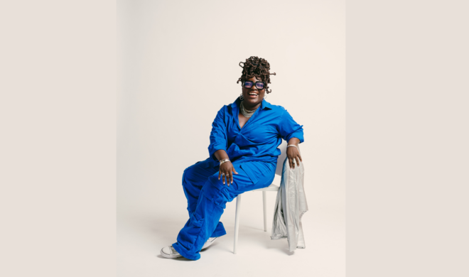 A photo portrait of Shahaddah Jack. She is Black. She is wearing bright blue coveralls, white sneakers, jewellery on her hands, neck, and ears. Her hair is in long curly dreads gathered at the top of her head. She is smiling widely. She is sitting in a white chair with a white jacket hung on it.