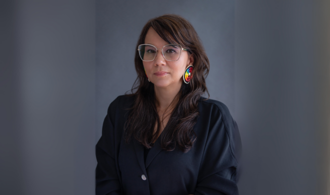 A photograph portrait of artist Tanya Lukin Linklater. She is an Indigenous woman with fair skin, and long brown hair. She has brown eyes and is wearing thin-framed glasses. She has a small smile on her face and is looking into the camera. She is wearing a black blazer.