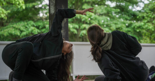 Two dancers, lisa nevada (left) and Emily Law (right), are on the floor, crouched in the middle of a movement. lisa has one knee on the floor, her body is tilted, and hands are open reaching towards the sky. Her head is tilted upwards similarly. Emily also has her head tilted upwards, with her body leaning sideways with her hand propping her body up for support. They are both wearing black clothing, joggers and hoodies. They are outside in a wooded green area, underneath a wooden gazebo.