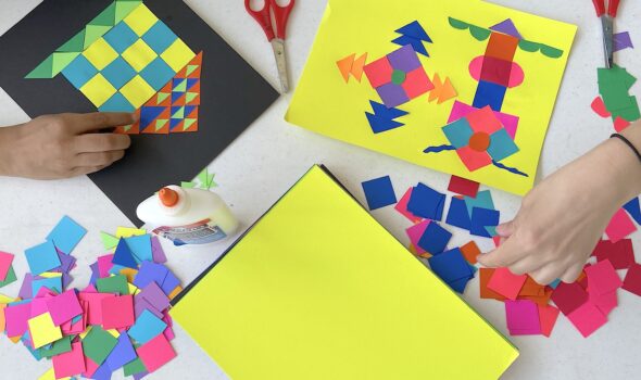 Taken from above, the photo is of different coloured construction paper cut into different shapes laid out in a table. They are glued together to make different symbols and art. Hands reach into the frame of the image. Scissors and glue are on the table.